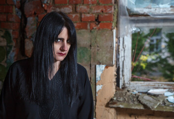 portrait of young goth woman standing near broken window of an abandoned house