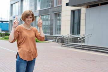 Obraz premium An adult red-haired woman with a serious face stands in the street with her fingers crossed. A gesture showing hope for something.