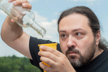 An adult man with a beard is trying to pour wine from a nearly empty bottle into a plastic cup. Man on a light background.