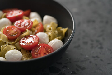 fettuccine pasta with pesto, mozzarella and cherry tomatoes in black bowl on concrete background closeup with copy space