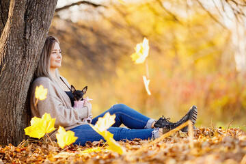 Beautiful girl jeans and warm sweater sits in autumn park with small terrier dog in her arms,...