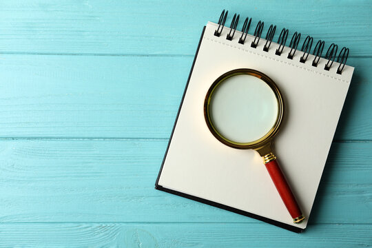 Top view of magnifier glass and empty notebook on light blue wooden background, space for text. Find keywords concept
