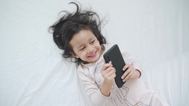 A 4 years old asian girl is smiling while she is using the smartphone on the bed in the morning time.