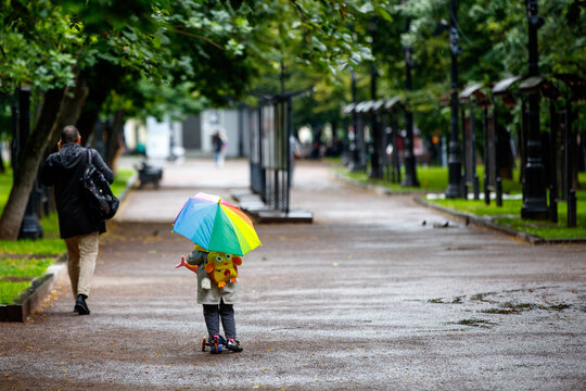 A Little Girl Walks In The Rain Along The Boulevard With A Multi-colored Broken Umbrella