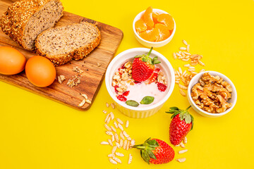 Healthy breakfast with granola, eggs, homemade bread,  and fruits on a wooden table, in white bowls, with a yellow background. 