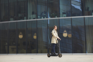 Graphic wide angle portrait of modern young businessman riding electric scooter across shot while commuting to work in downtown with urban city buildings in background, copy space © Seventyfour