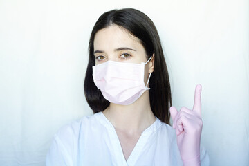 A woman in gloves and a protective mask stands on a light background in a white jacket. The concept of suppression of coronavirus