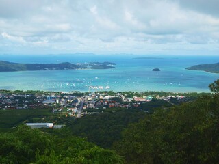 Fototapeta na wymiar Panoramic view from the top to the bottom to the coastal town. Sea bay. Turquoise water and sky with gray cumulus clouds. Seascape. Group of islands on the horizon. Harbor with boats. Phuket, Thailand