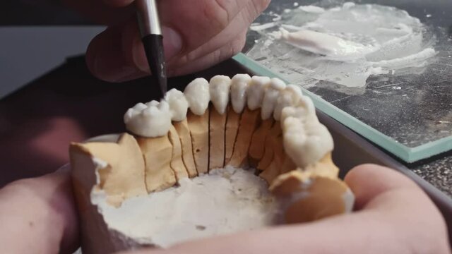 Close up view of dental technician draws white ceramics glaze on prosthesis of teeth in workplace in laboratory of implants