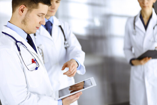 Group of doctors are checking medical names on a computer tablet, with a nurse with a clipboard on the background, standing together in a hospital office. Physicians ready to examine and help patients