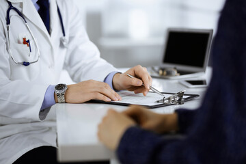 Fototapeta premium A doctor is talking to his patient, while sitting together at the desk in the cabinet in a hospital. Physician using clipboard for filling up medication history records. Perfect medical service in