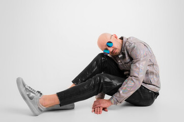 Stylish caucasian man without hair in grey shirt, black leather trousers and grey trainers with black sunglasses, picture isolated on white background