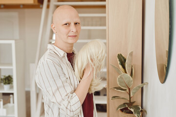 Warm-toned portrait of bald adult woman looking at camera holding wig while standing by mirror in...