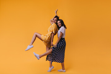 Funny tanned girl in black skirt fooling around with best friend. Studio portrait of laughing amazing ladies dancing on orange background.