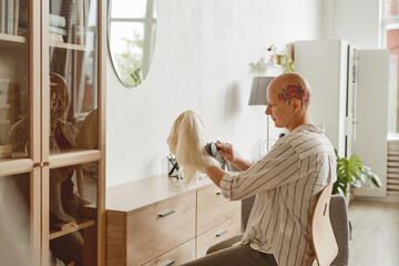 Side view portrait of modern bald woman brushing wig while sitting by mirror in home interior, alopecia and cancer awareness, copy space - obrazy, fototapety, plakaty