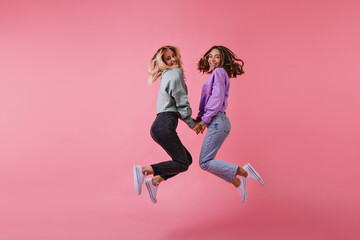 Studio portrait of blithesome best friends holding hands on pink background. Charming sisters in trendy pants jumping and laughing.