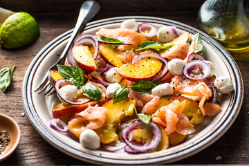 Refreshing summer salad with smoked salmon, peaches, mozzarella, red onion and basil. Wooden background.