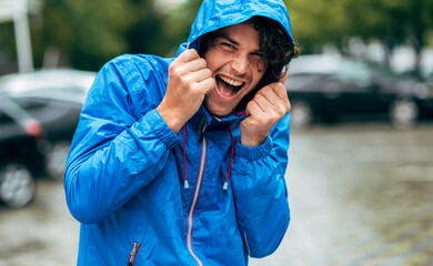 Happy man laughing and wearing blue raincoat during the rain outside. Handsome male in blue raincoat enjoying the rain in the city street. Male has joyful expression in rainy weather.