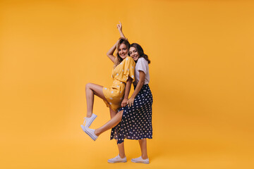 Blissful brunette girl in long skirt posing with her sister. Indoor shot of spectacular female friends isolated on yellow background.