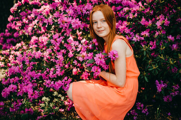 Strange young girl with long red hair, pretty face in a summer orange dress have a good time in the garden