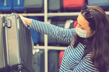 A young woman in a disposable mask choosing a suitcase for the upcoming vacation after the end of...