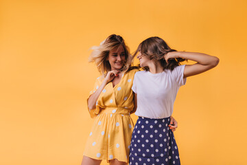 Blithesome young woman in yellow outfit looking at friend with smile. Joyful caucasian sisters chilling on orange background.