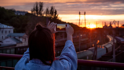 A young woman photographs a beautiful sunset from a bridge.