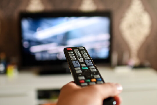 Close up of television remote control in the hand in front of TV set. Selective focus. Low DOF
