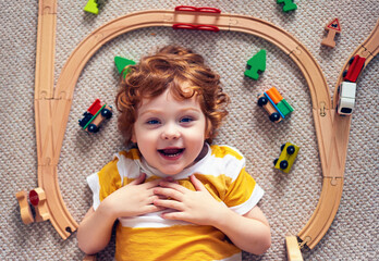 adorable happy redhead baby boy laying on the carpet among railway tracks and trains, wooden toys