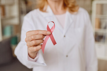 Warm-toned close up of unrecognizable female doctor holding pink ribbon as symbol of breast cancer...