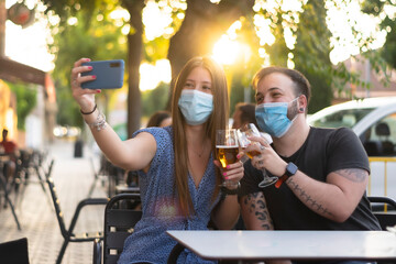 Spain Madrid. Caucasian couple toasting with beer wearing sanitary mask taking a selfie. Reopen...