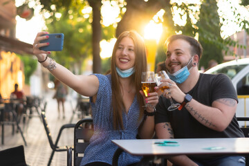 Spain Madrid. Caucasian couple toasting with beer wearing sanitary mask taking a selfie. Reopen pubs and club after quarentine coronavirus Covid-19. Man and woman drinking beer. Cheers. 

