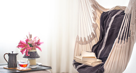 A boho-style hammock chair with a stack of books. Wooden tray with teapot and Cup of tea and beautiful Lily flowers. Cozy interior. The concept of rest and home comfort.