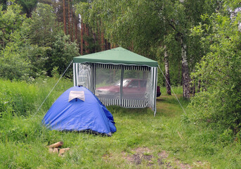 Blue small tourist tent with a tent and a bard car in the woods. Comfortable outdoor recreation.  Camping tent stand in the shade of a pine forest.