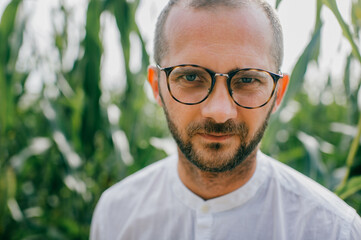 Portrait of a beautiful caucasian male with short dadrk hair, black bristles on his face with glasses and a white shirt poses for the camera in cornfield