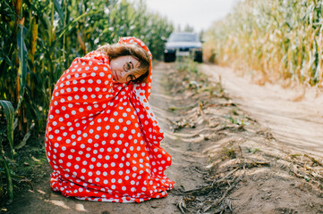 Beautiful caucasian female with short dark wavy hair in glasses wrapped in red and white coverlet have a lot of fun in cornfield in summer morning