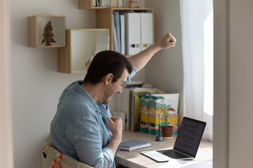 Overjoyed young man sit at desk at home look at laptop screen celebrate online win or victory, excited male triumph feel euphoric get good news or promotion letter on computer, success concept