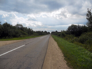 Rural road in warm summer. Green grass and blue sky with clouds.