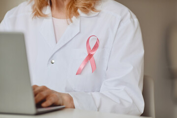 Close up background of pink ribbon pinned on white lab coat of female doctor using laptop, breast...