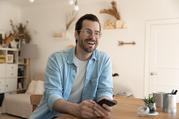 Happy millennial Caucasian guy in glasses sit at desk at home have fun talking chatting with friend, overjoyed young man smile laugh engaged in pleasant conversation, communication concept