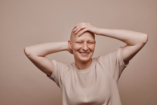 Warm-toned waist up portrait of carefree bald woman touching shaved head and smiling while posing against minimal beige background in studio, alopecia and cancer awareness, copy space