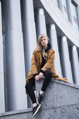 Young attractive teenager model with blonde long hair wearing orange trench and black hoodie sitting with background of huge columns in big city. Lifestyle and fashion concept.