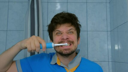 Funny man is brushing teeth usin electric toothbrush and making cheerful faces. Dental Oral hygiene concept. Young guy is looking at camera as at mirror. Happy person doing his morning routine.
