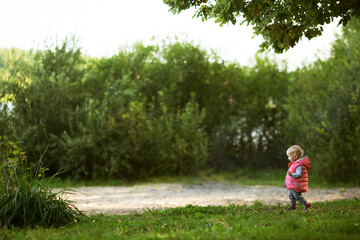 Naklejka premium Baby girl with blond hair wearing pink waistcoat walking in green park exploring the big and interesting world