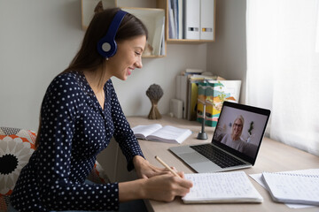 Smiling girl in earphones sit at desk at home look at laptop screen study distant with teacher,...