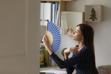 Overheated young Caucasian woman sit at desk at home wave with hand fan suffer from hot weather no...