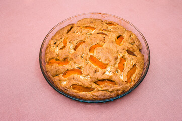 A large, round baked apple and apricot pie stands in a glass form on a table with a tablecloth.