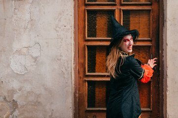 Charming female in witch costume poses outside in front of the big old door in black and red dress