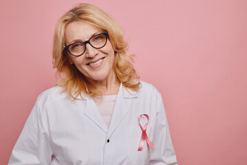 Waist up portrait of mature female doctor with pink ribbon on white coat smiling at camera while posing against pink background in studio , copy space - Powered by Adobe