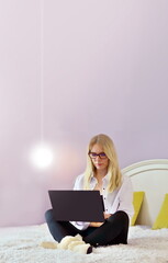 Happy casual beautiful woman working on a laptop sitting on the bed in the house.